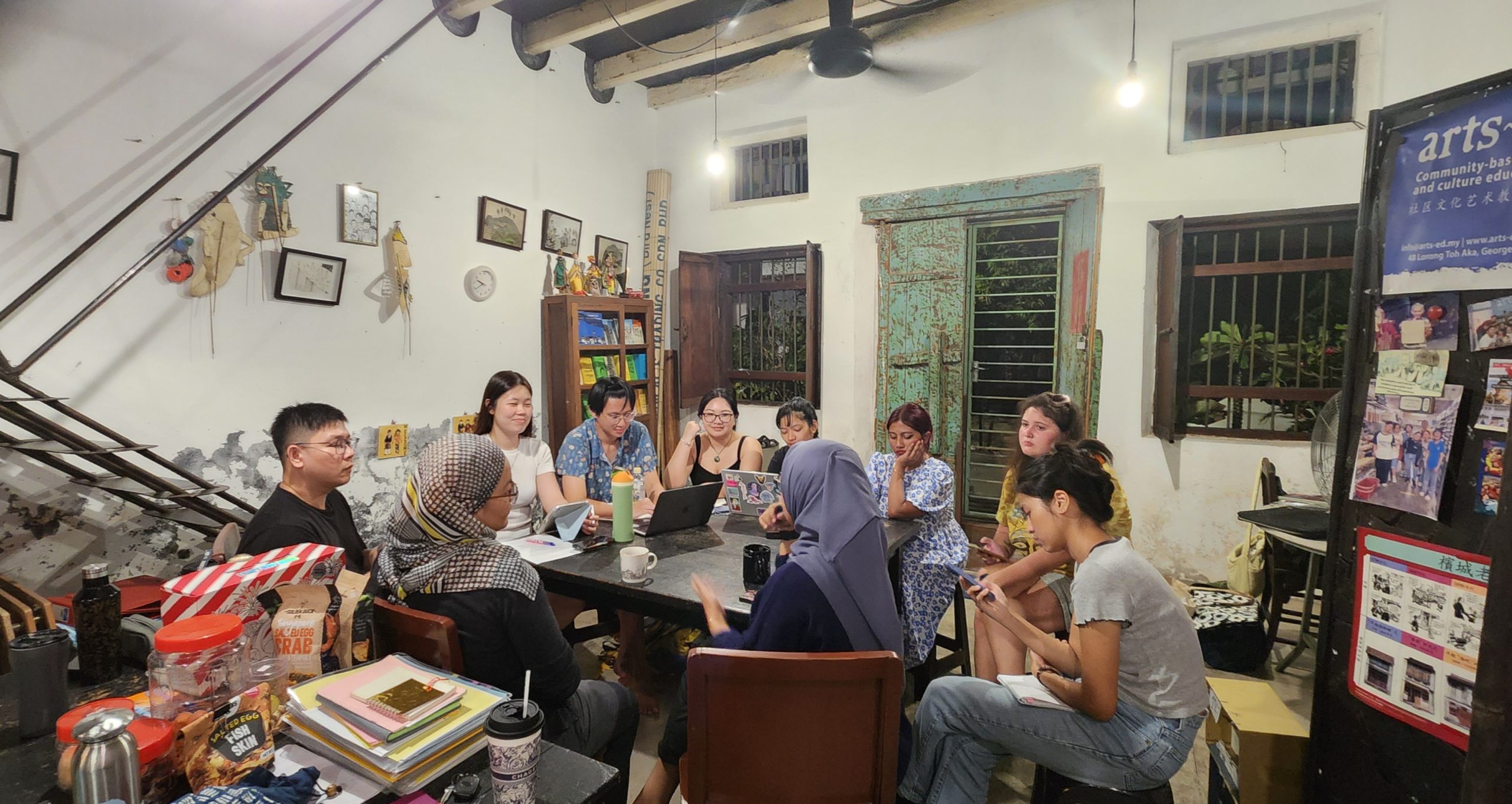 People seated around a large table in a rustic community space, engaged in discussion with notebooks, laptops, snacks and decorations.