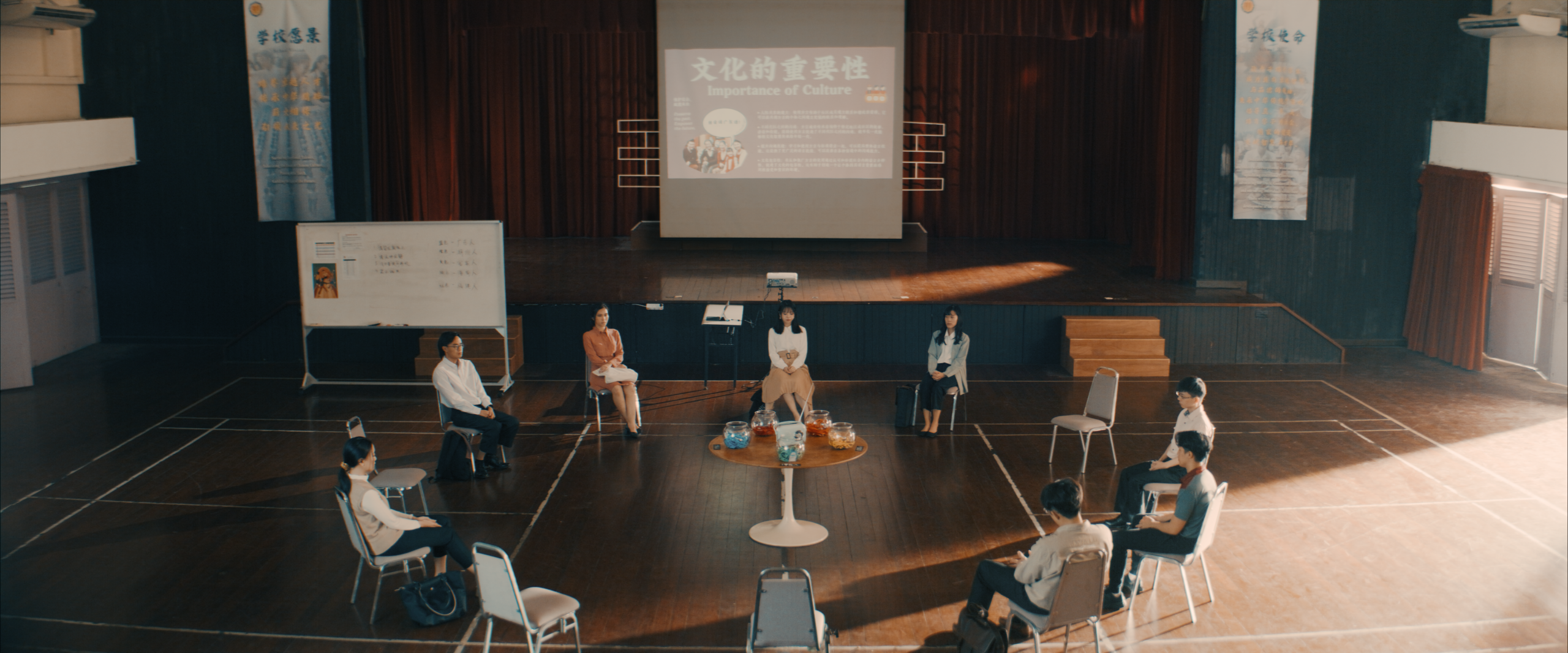 Group seated in a circle on a wooden floor in a school hall, viewing a projector screen titled “Importance of Culture” in English and Chinese.