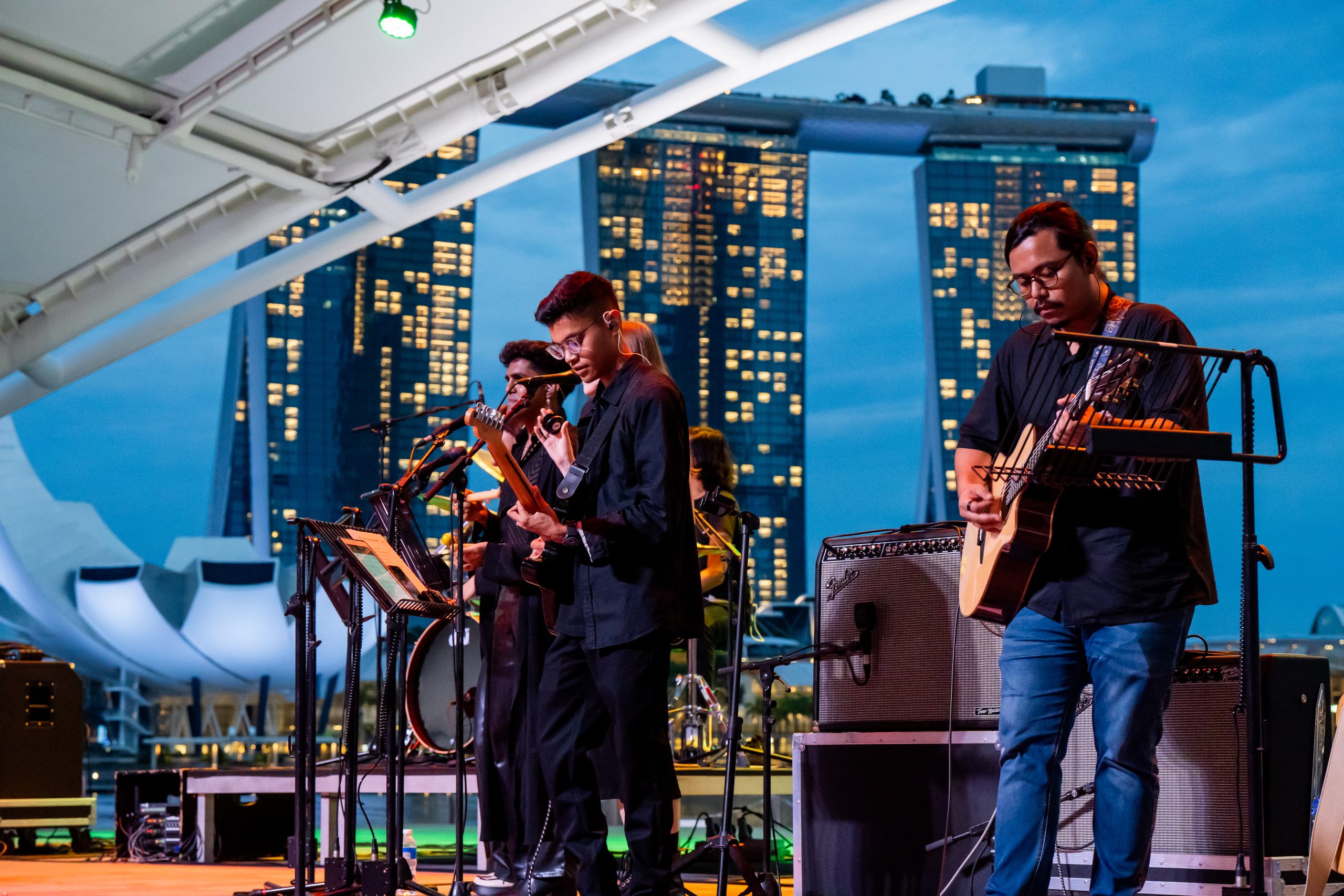 Student band performs at dusk under a canopy, with guitars and microphones, against Marina Bay Sands and ArtScience Museum skyline.
