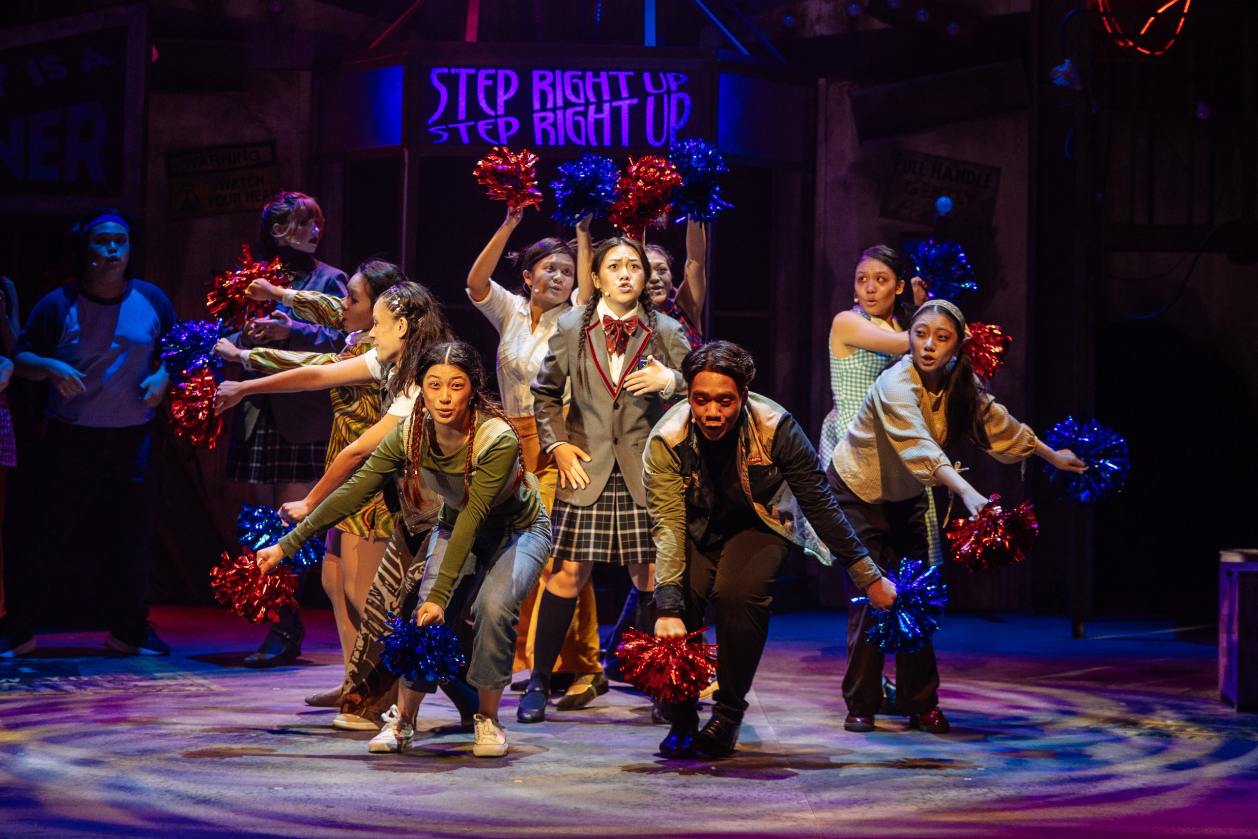 Performers on stage mid-action with pom-poms, school uniforms and a glowing “Step Right Up” sign under theatrical lighting.