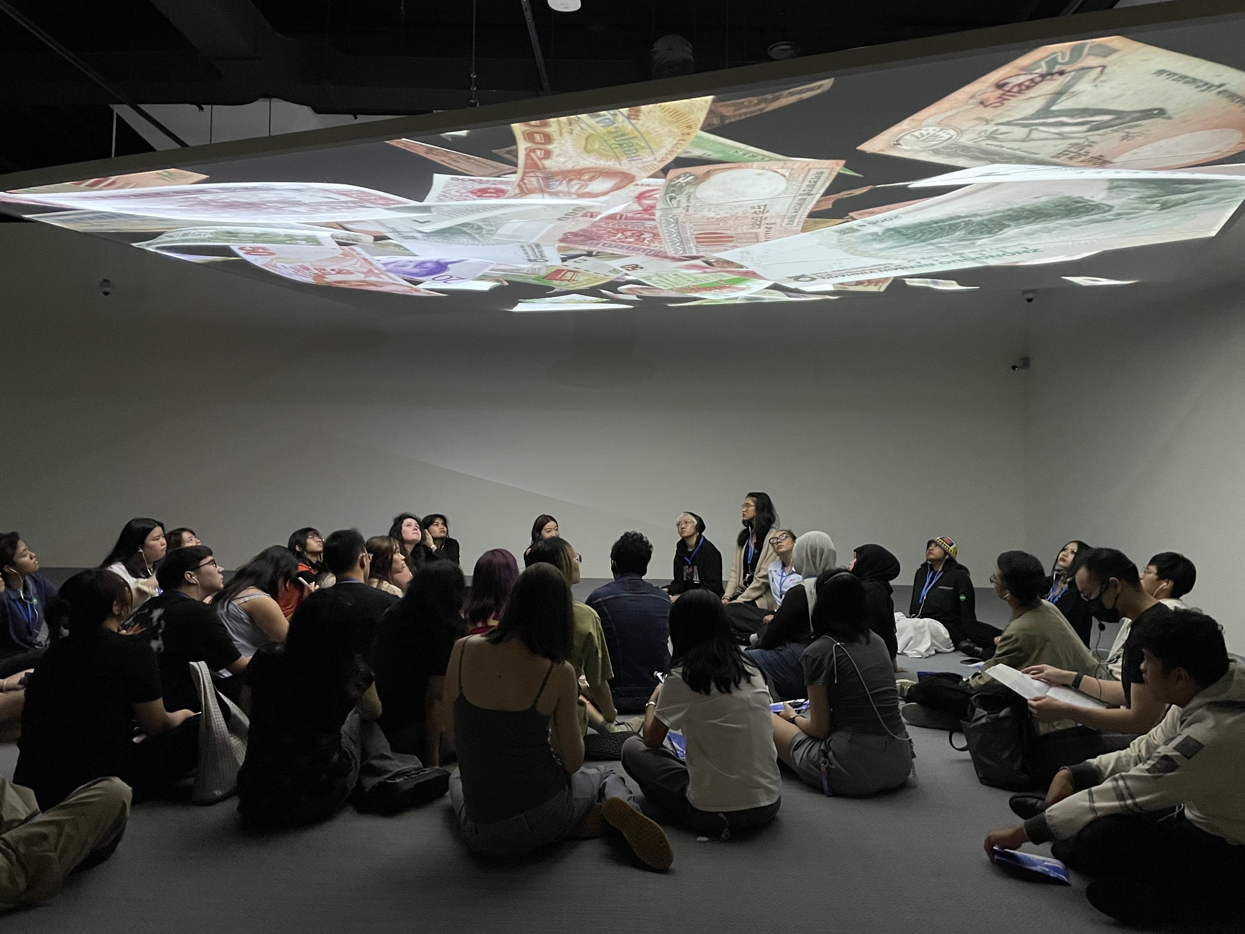 Students seated in a darkened gallery space, engaged in a discussion beneath a ceiling projection of international currency notes.