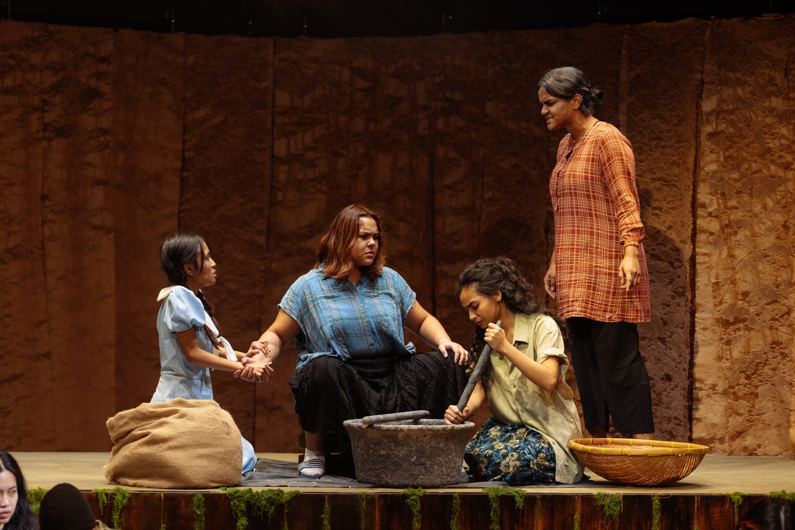 Four women in traditional Malay attire perform on stage with props including a child, stone grinder and rustic set evoking a village scene.