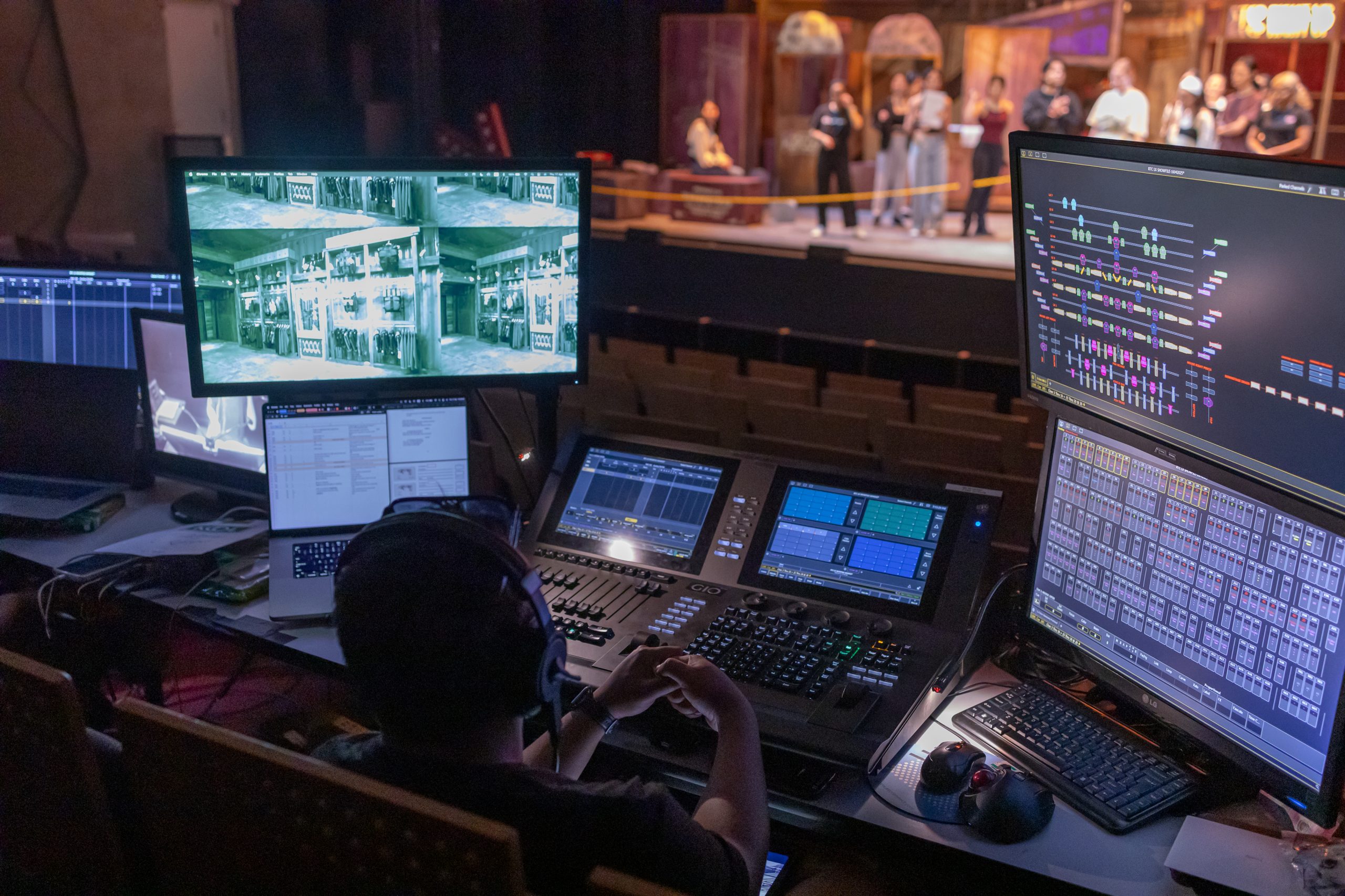 Stage technician operates control boards with screens showing lighting plots and camera feeds, as performers rehearse in the background.