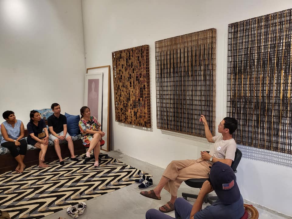 Artist seated by large woven wall art leads a barefoot audience sitting on a patterned rug during an intimate gallery talk in a white room.