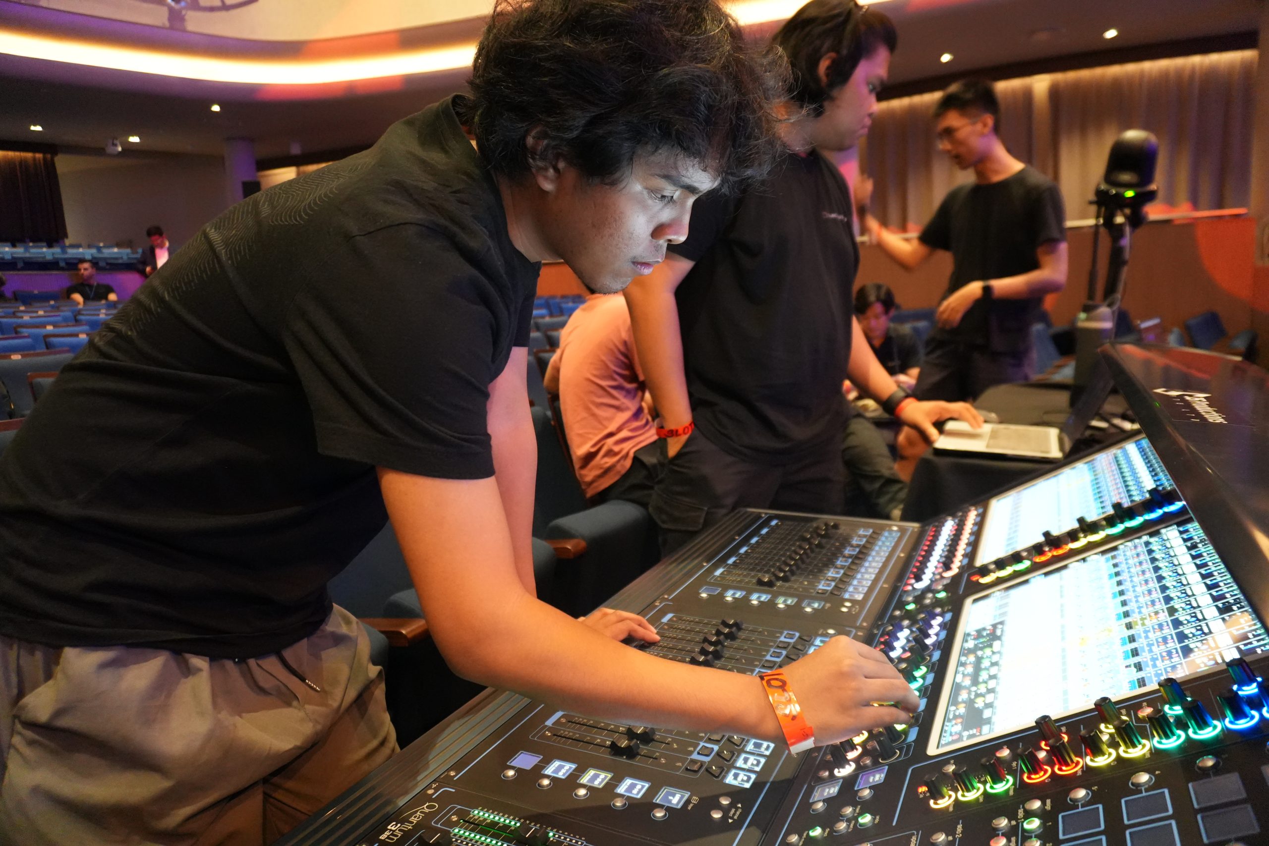 Student operates a digital sound mixing console in a dim auditorium while others stand behind him in discussion during a live session.