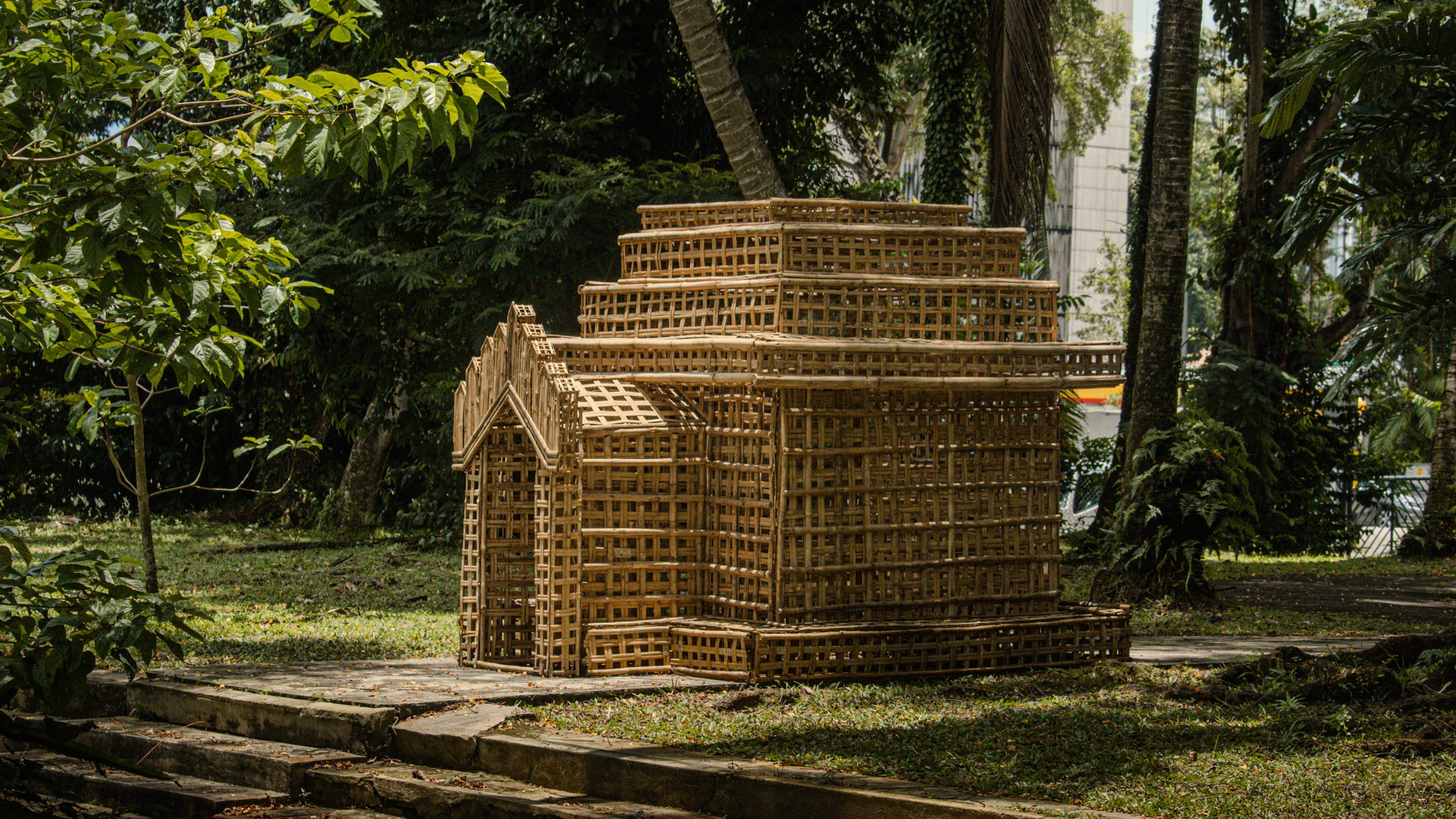 Miniature architectural structure woven from bamboo strips sits outdoors in a tropical garden with dappled sunlight filtering through trees.
