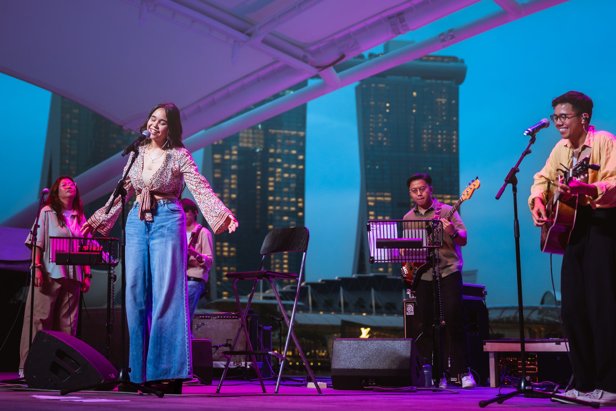 Live band performing on an outdoor stage at dusk with Marina Bay Sands and ArtScience Museum visible in the background.