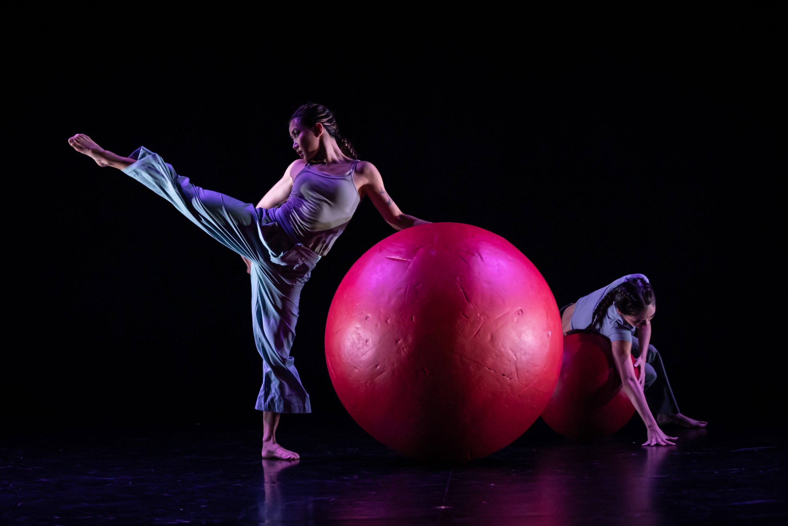 Two performers on a dark stage interacting with large red exercise balls during a contemporary dance piece.