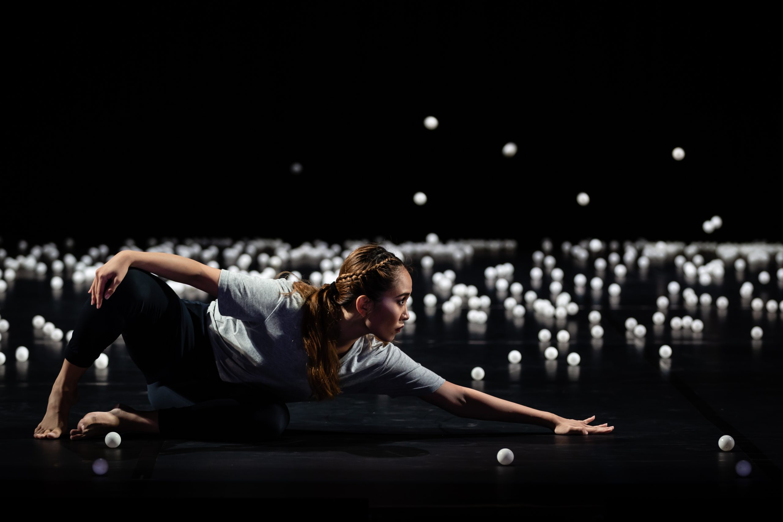 Dancer in grey shirt and black pants performs barefoot on a dark stage scattered with white balls, arm extended in a low pose.