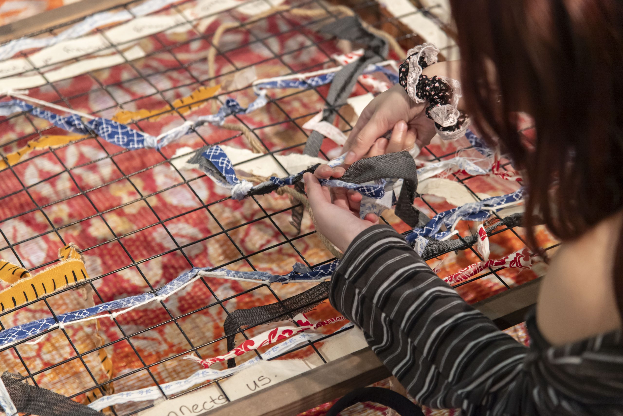 Close-up of hands weaving colourful fabric strips through a metal grid, part of a textile installation over a red patterned backdrop.