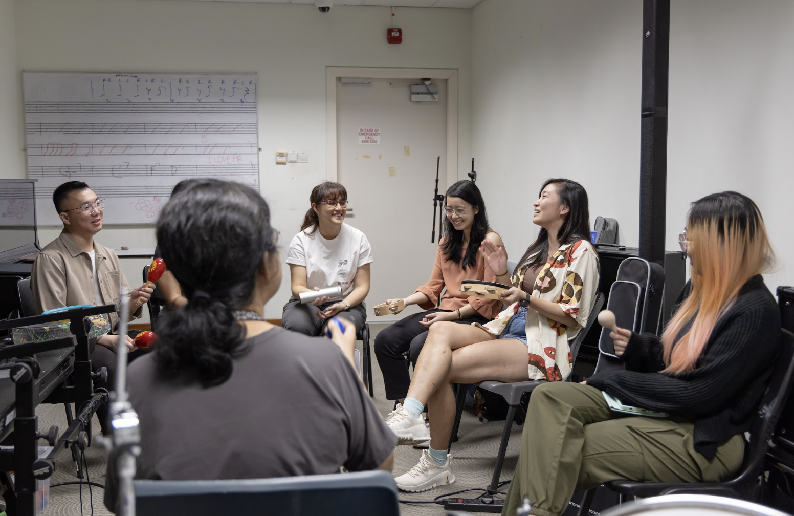 Students seated in a circle play tambourines and maracas in a music classroom, with a whiteboard displaying music notation behind them.