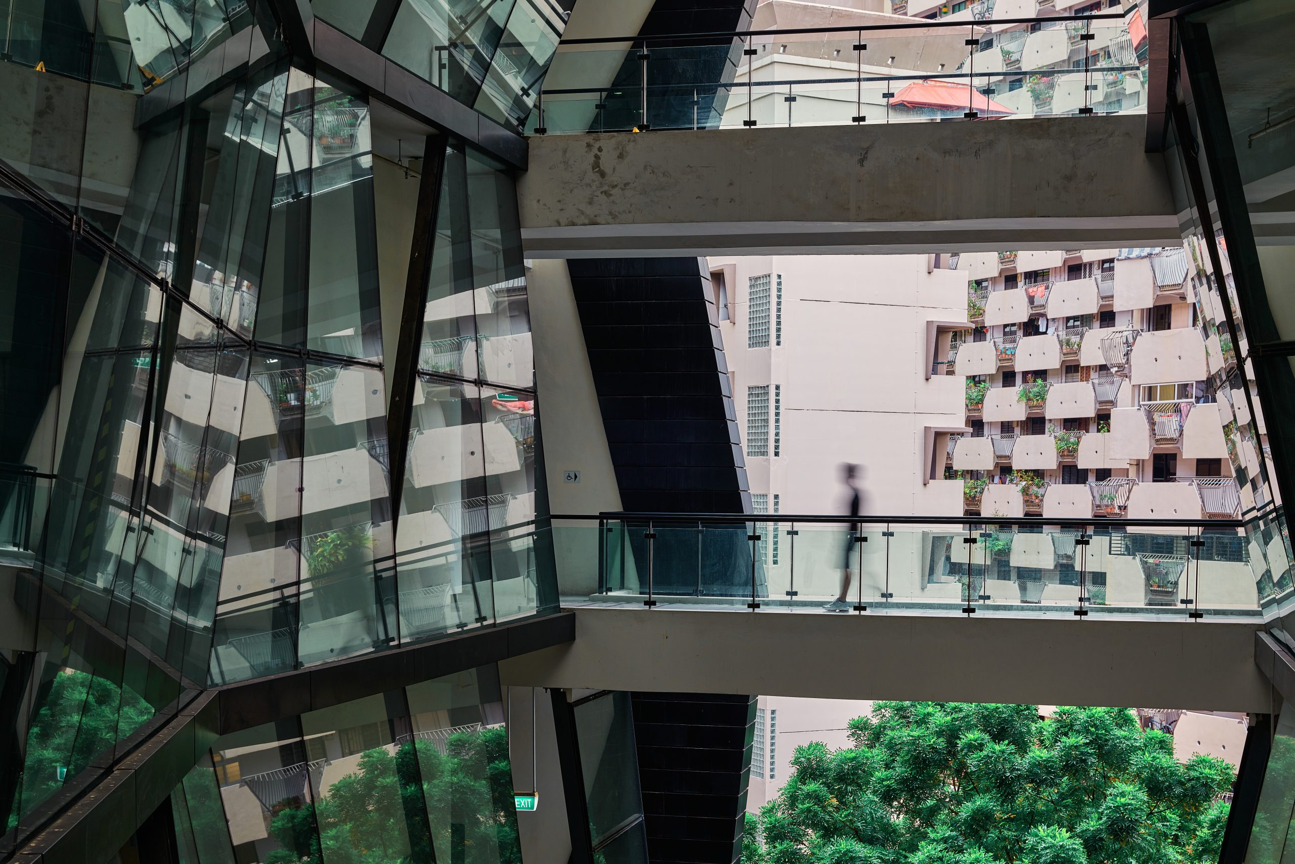 Blurred silhouette walks across glass bridge inside LASALLE McNally Campus, with angular architecture reflecting nearby flats and greenery.