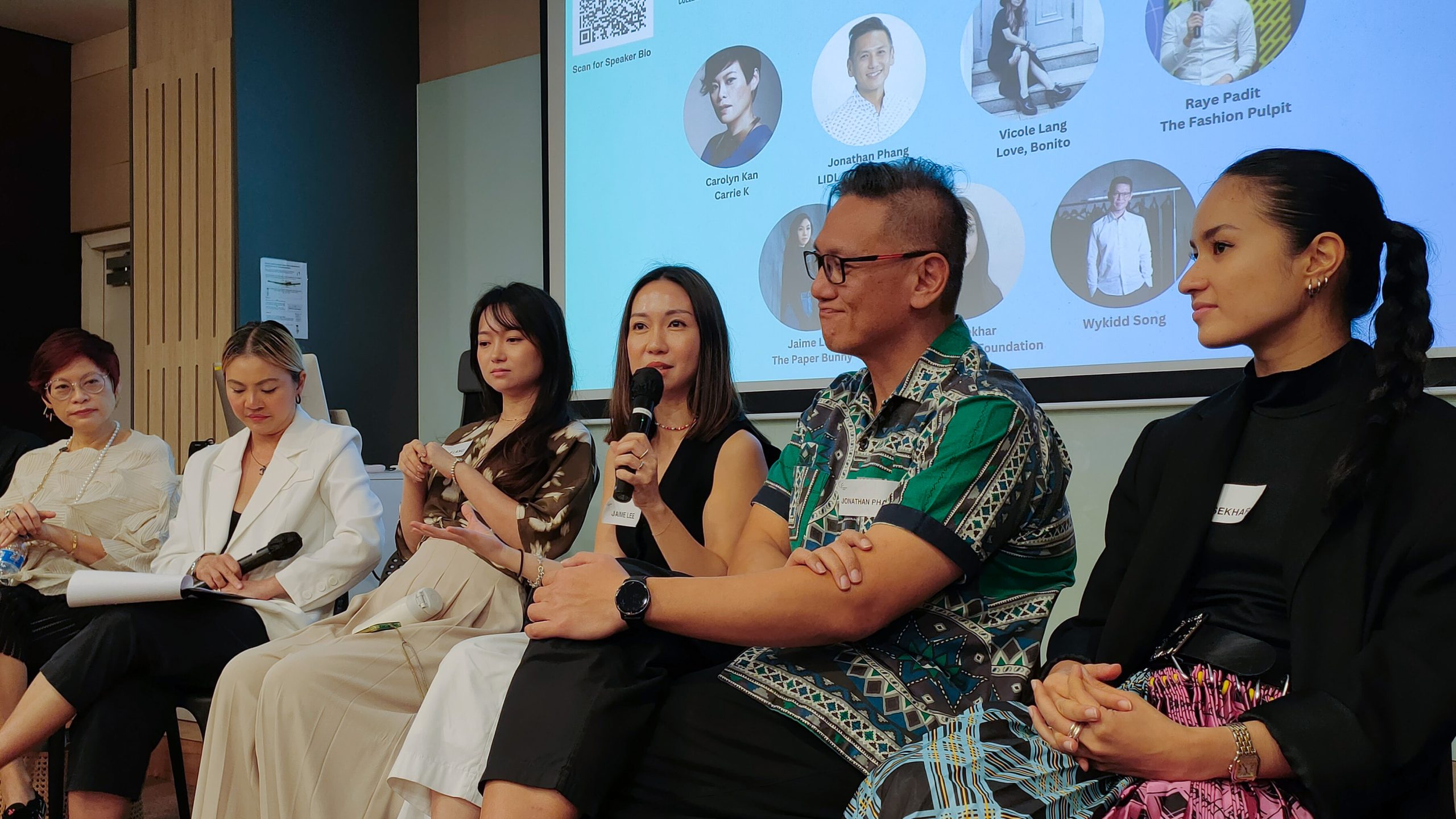 A seated panel of speakers in mid-discussion, with one woman holding a microphone, in front of a screen displaying headshots and names.