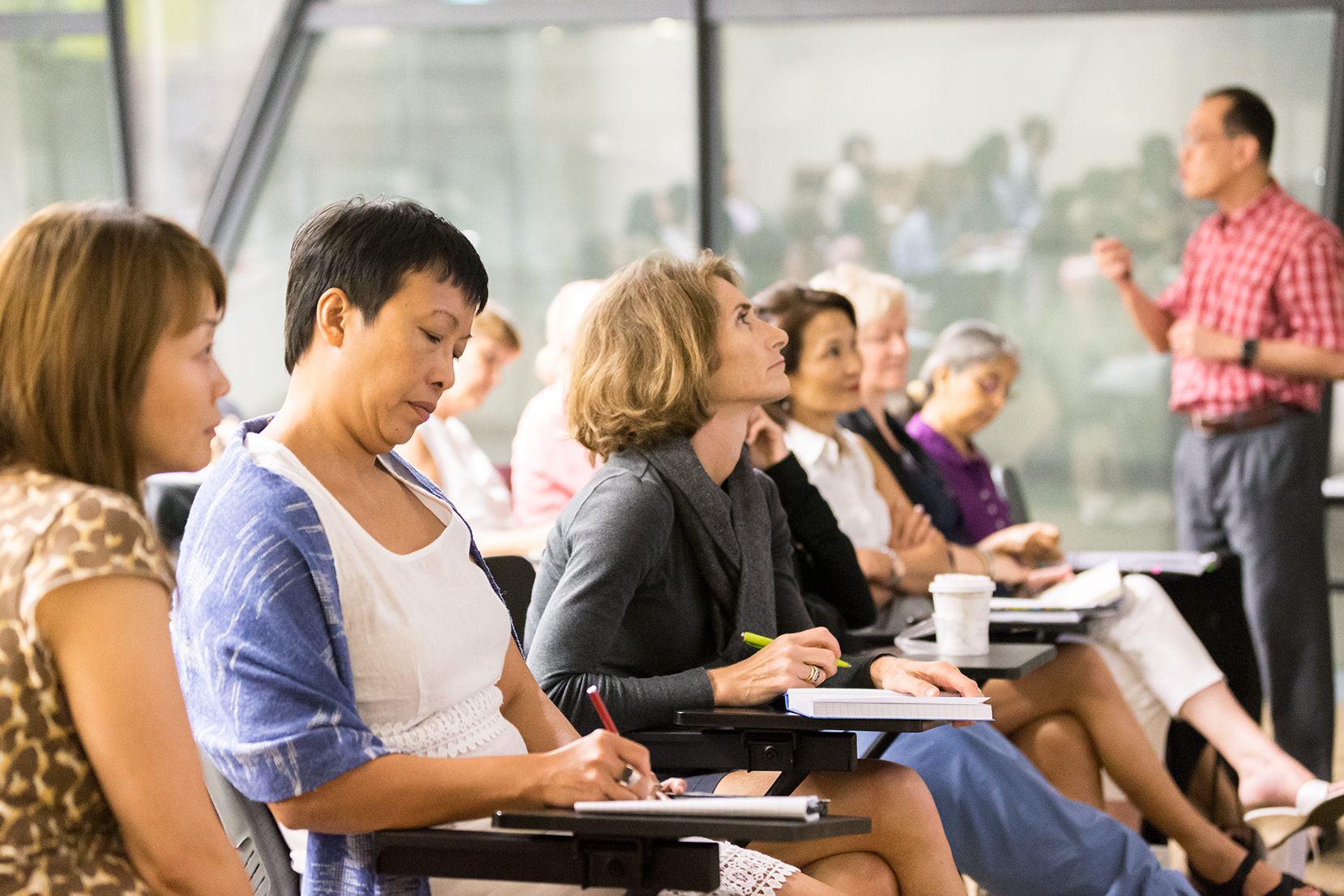Adult learners seated in a classroom, attentively listening and taking notes during a lecture by a male speaker in a red checkered shirt.