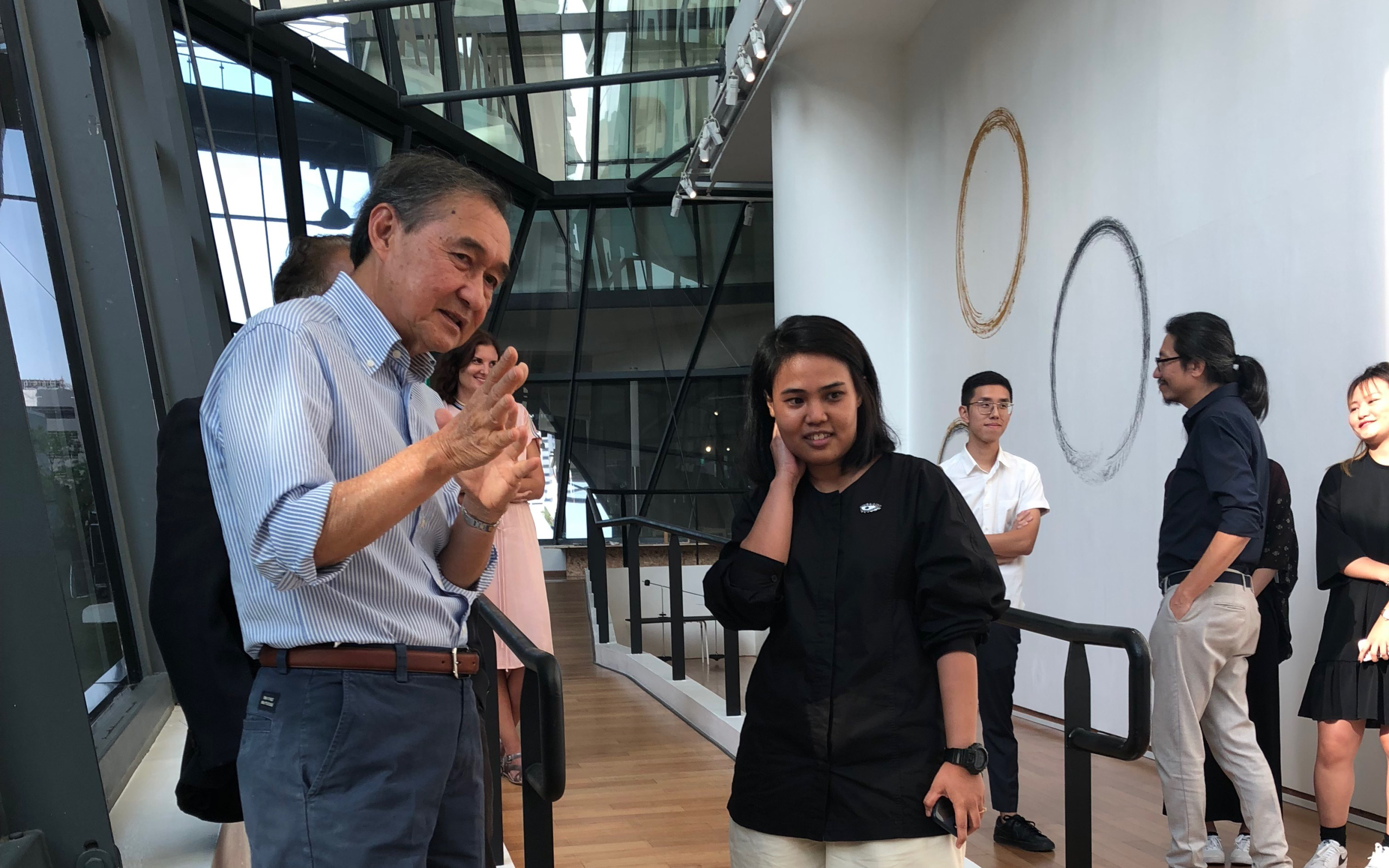 LASALLE visitors and students engaged in conversation inside a light-filled gallery walkway with circular wall art and glass panels.
