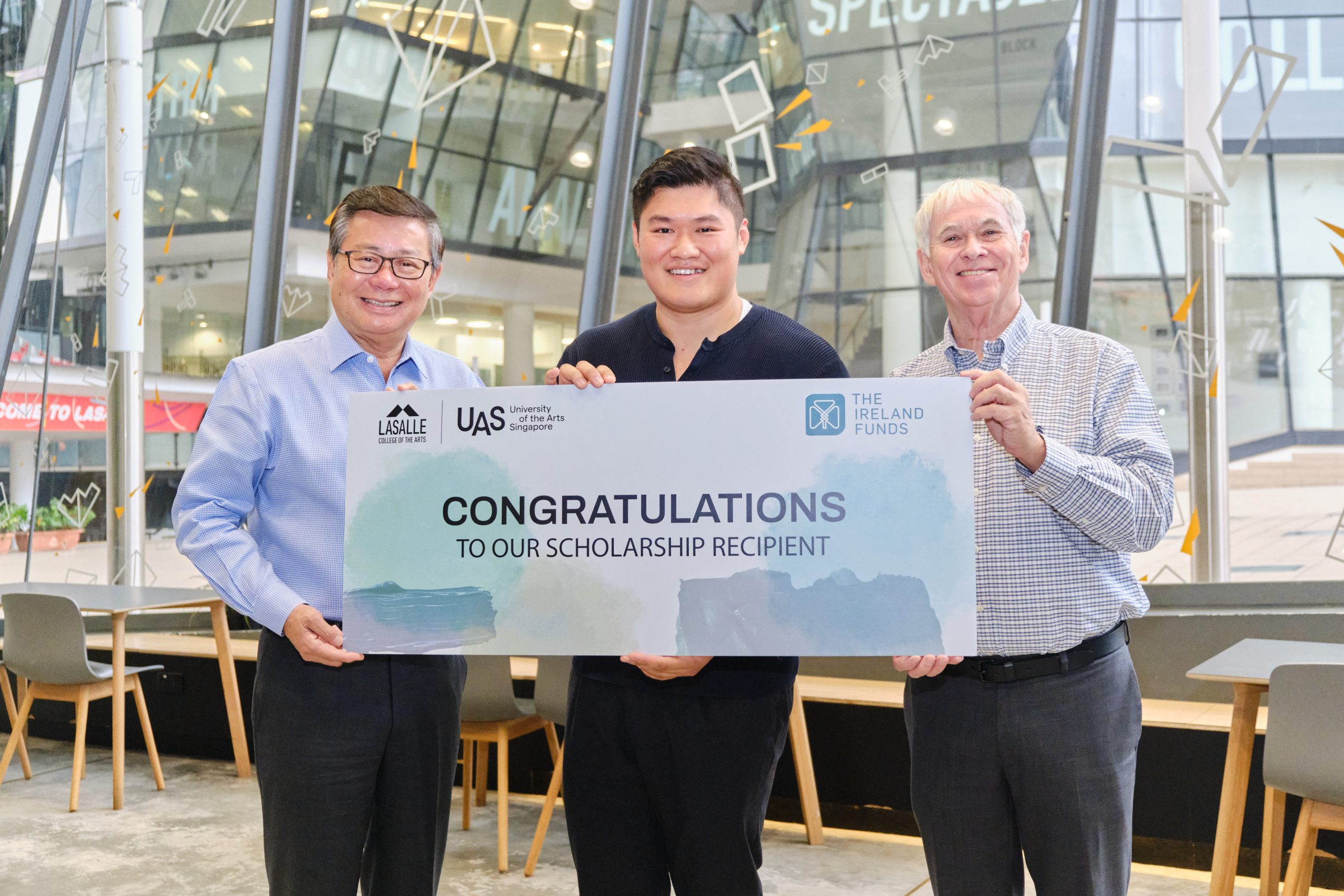 Three men smiling and holding a scholarship award banner at LASALLE, with UAS and The Ireland Funds logos.