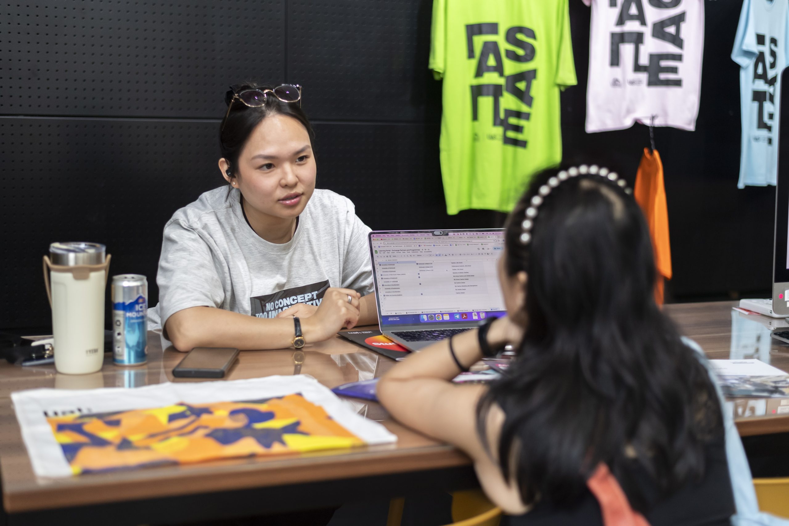 Two students in conversation at a LASALLE booth, with open laptops and colourful printed materials on the table, and graphic LASALLE T-shirts displayed behind them.