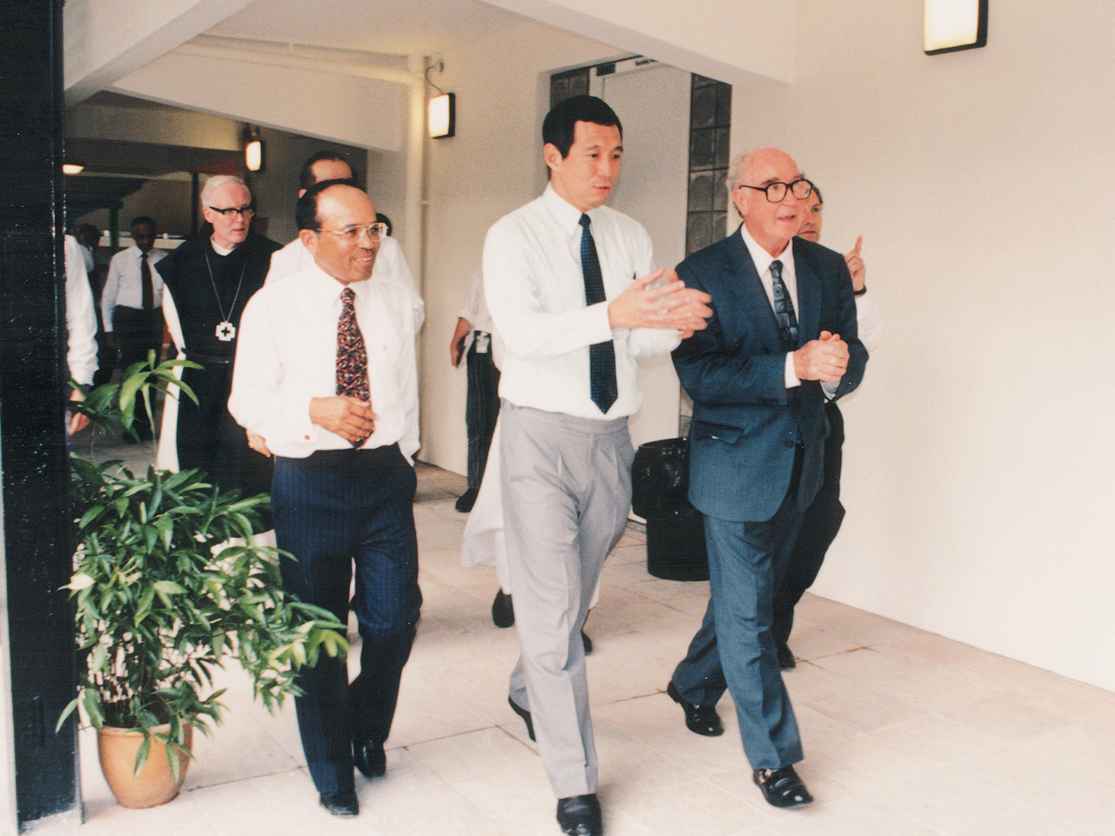 Vintage photography of a group of men in formal attire, including a central figure gesturing while walking, touring a LASALLE campus corridor with potted plants and soft lighting.