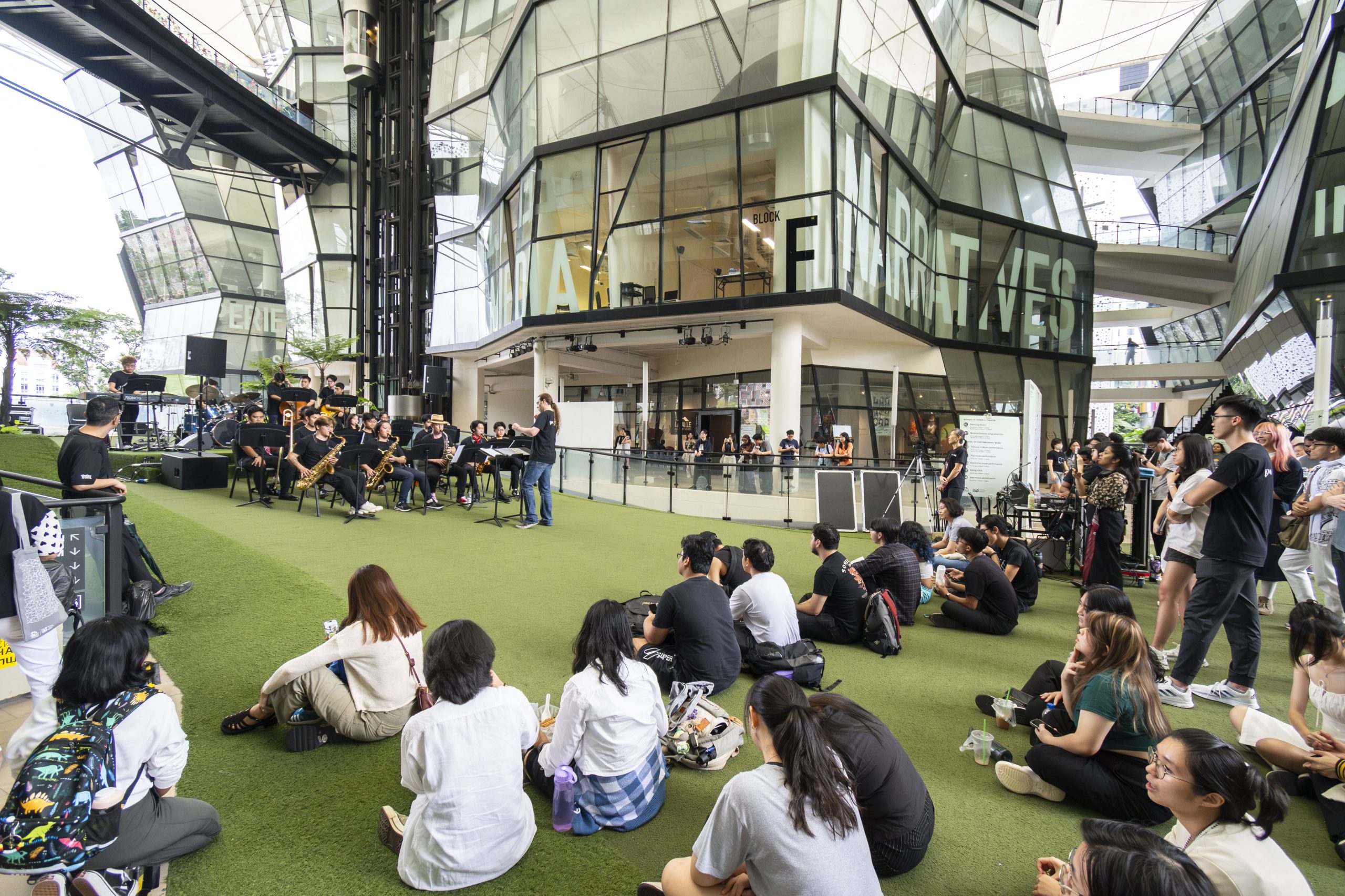 Large group of students seated on the McNally Campus green, watching a live jazz band performance surrounded by LASALLE’s glass architecture.