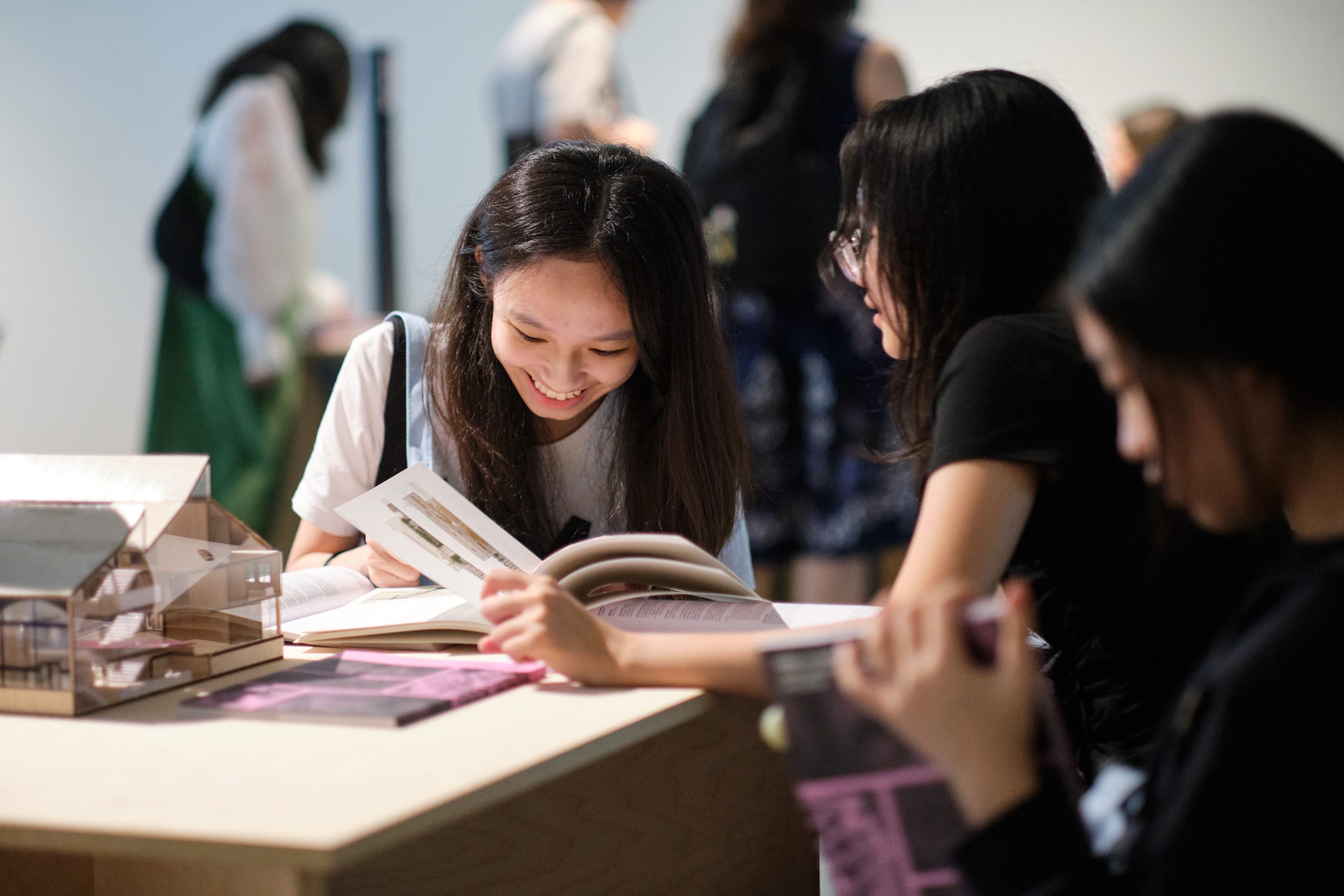 Three students read design books at a table with architectural models, smiling and discussing pages in a well-lit gallery space.