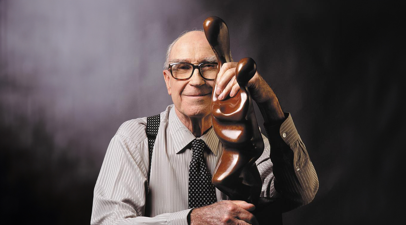 Brother Joseph McNally smiling while holding an abstract wooden sculpture against a dark studio backdrop.