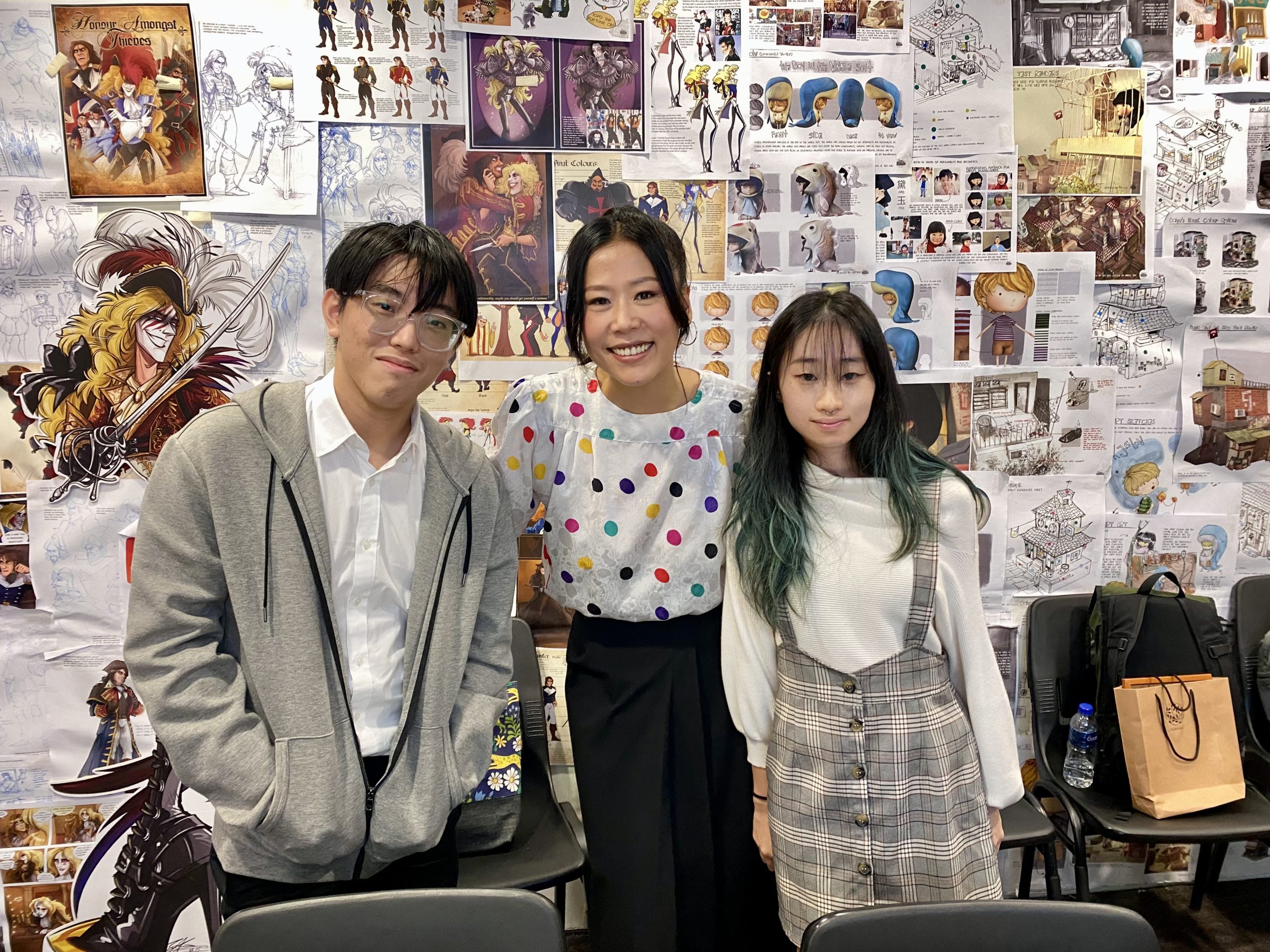 Three people smiling in front of a colourful LASALLE student concept art wall filled with character sketches, digital illustrations and visual development boards.