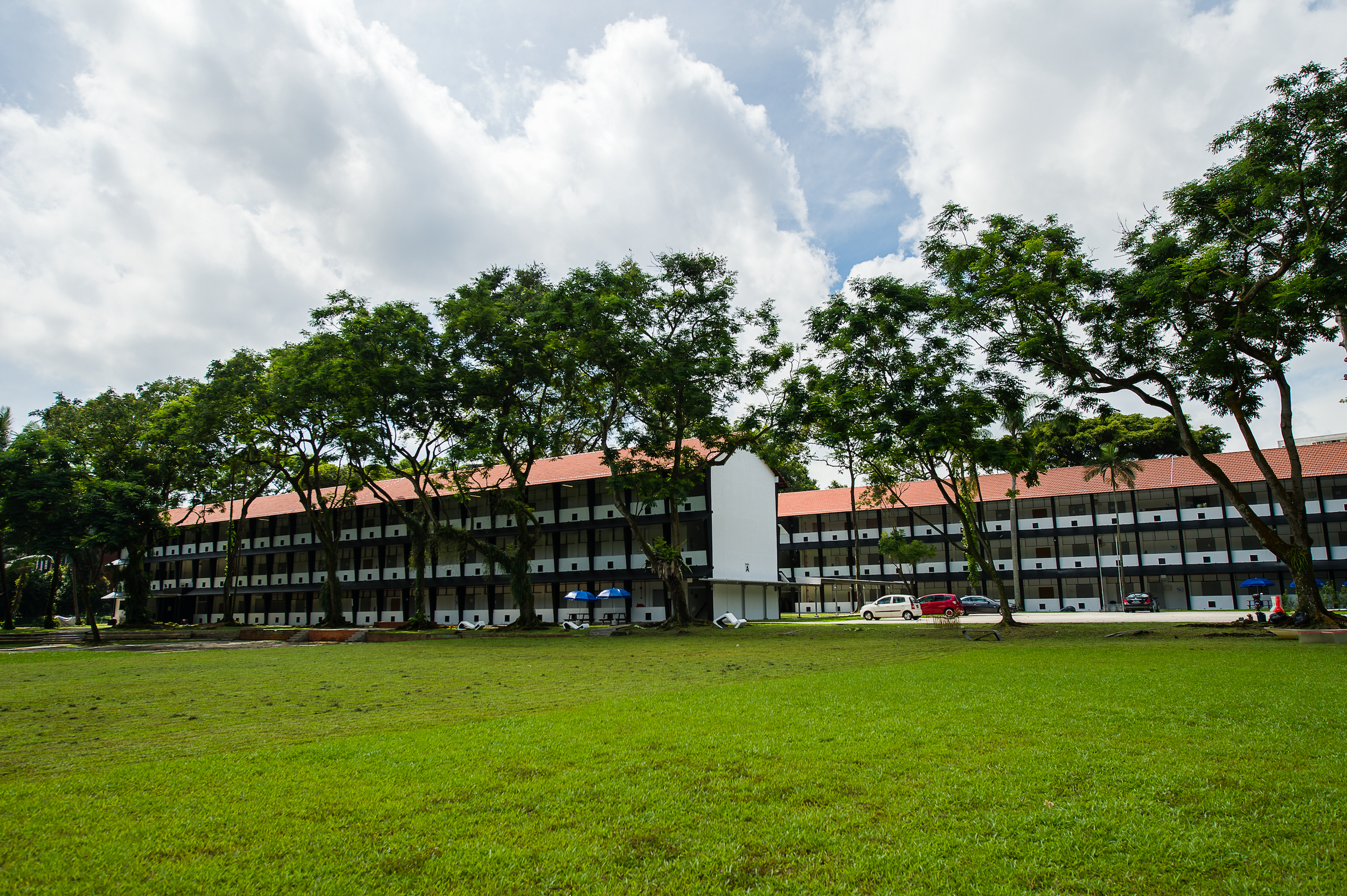 Exterior view of a long three-storey building with red roofs and shaded walkways, set behind a wide green lawn and tall trees.