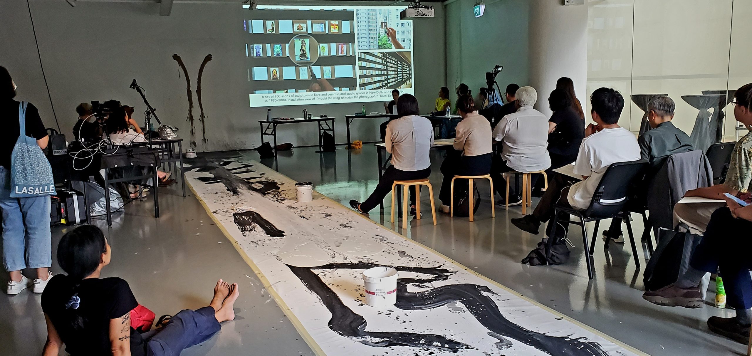 Audience at a LASALLE workshop watch a presentation as a long painted canvas stretches across the floor in front of them.