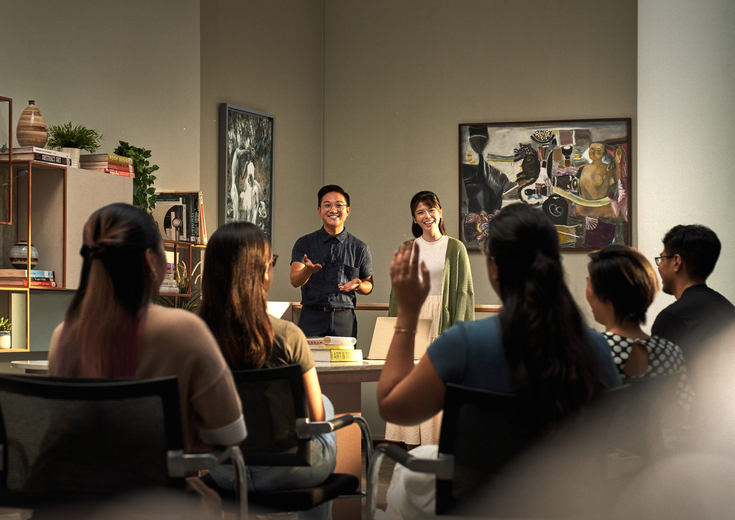 Two presenters lead a classroom discussion with students seated in front of them, surrounded by bookshelves and framed paintings.