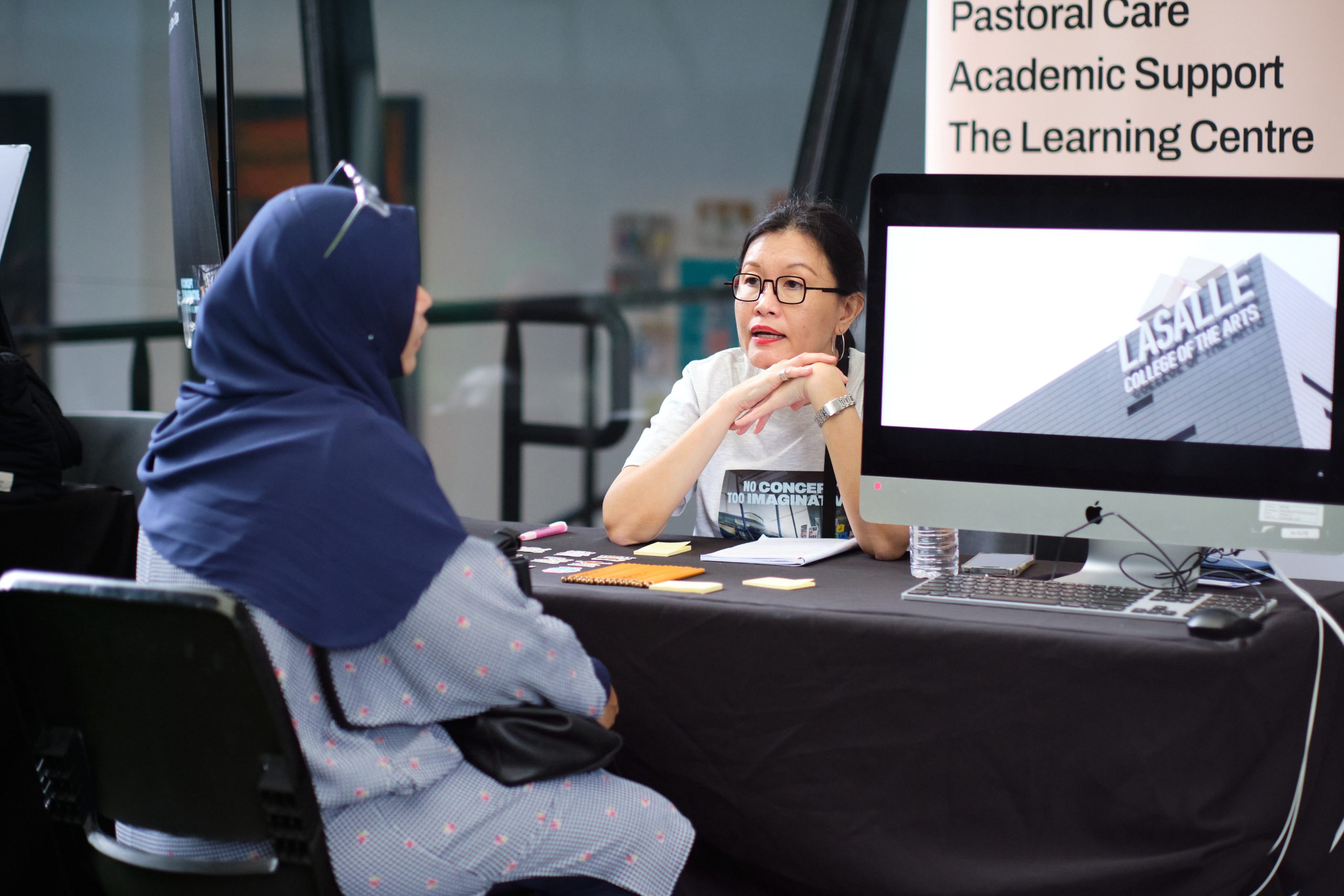 LASALLE staff member speaking with a prospective student at a support services booth, with a screen displaying the college's façade.