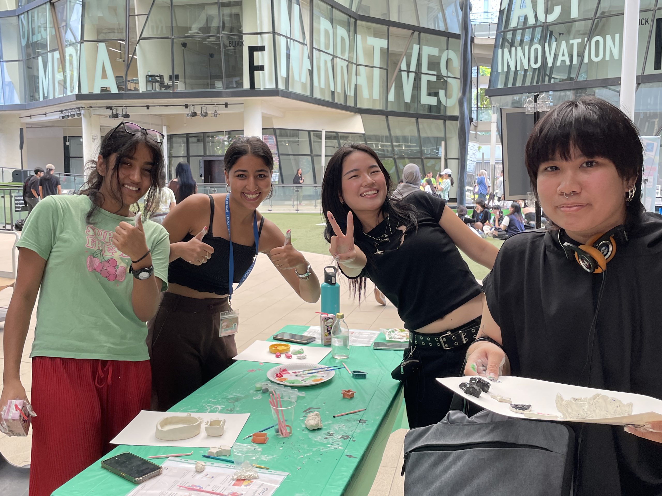 Four students at LASALLE smile and pose with clay art pieces on a green table, set against McNally Campus' Block H at Campus Green.