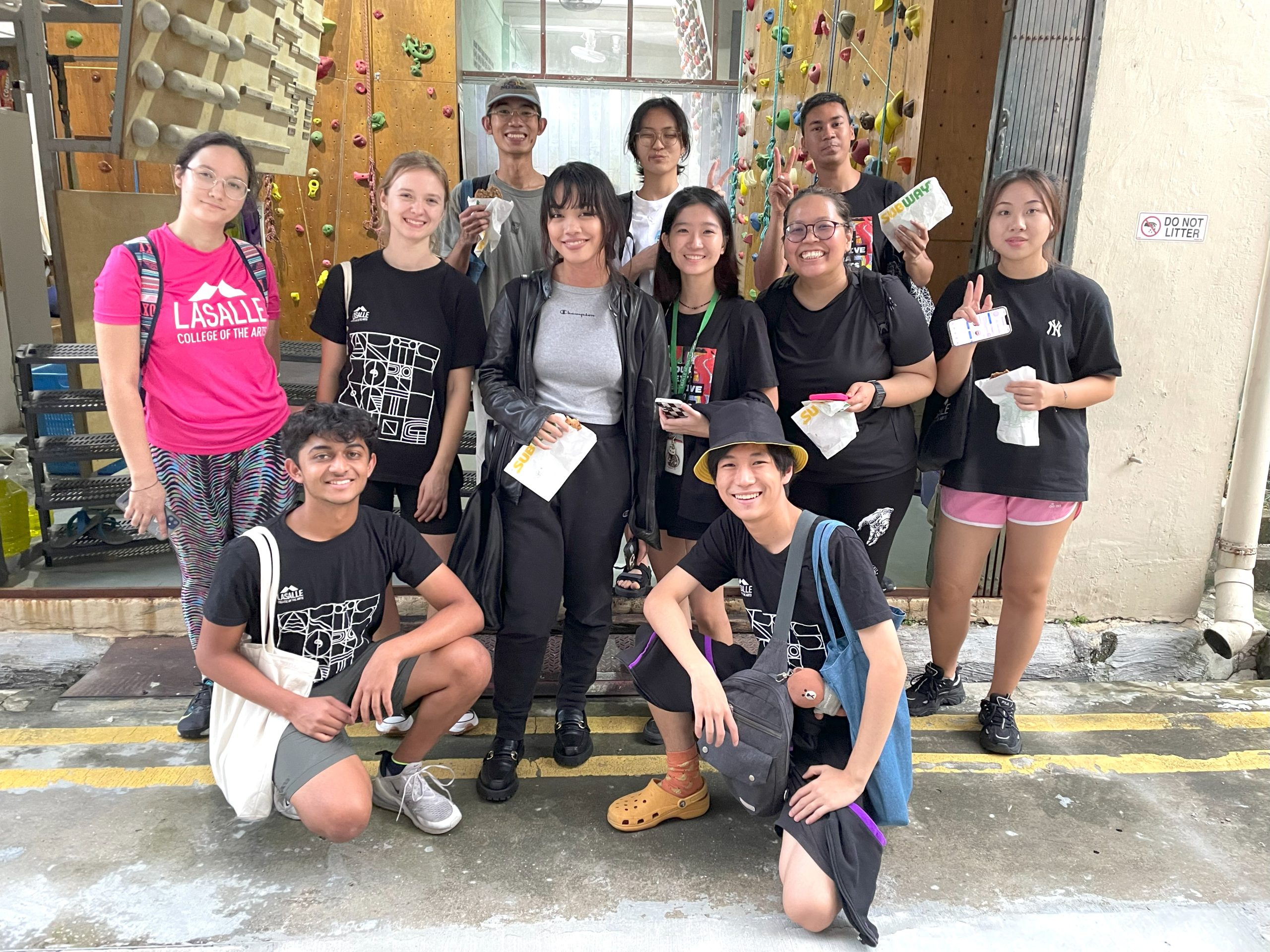 Group of LASALLE students posing outside a climbing gym, some holding Subway sandwiches, with a bouldering wall in the background.