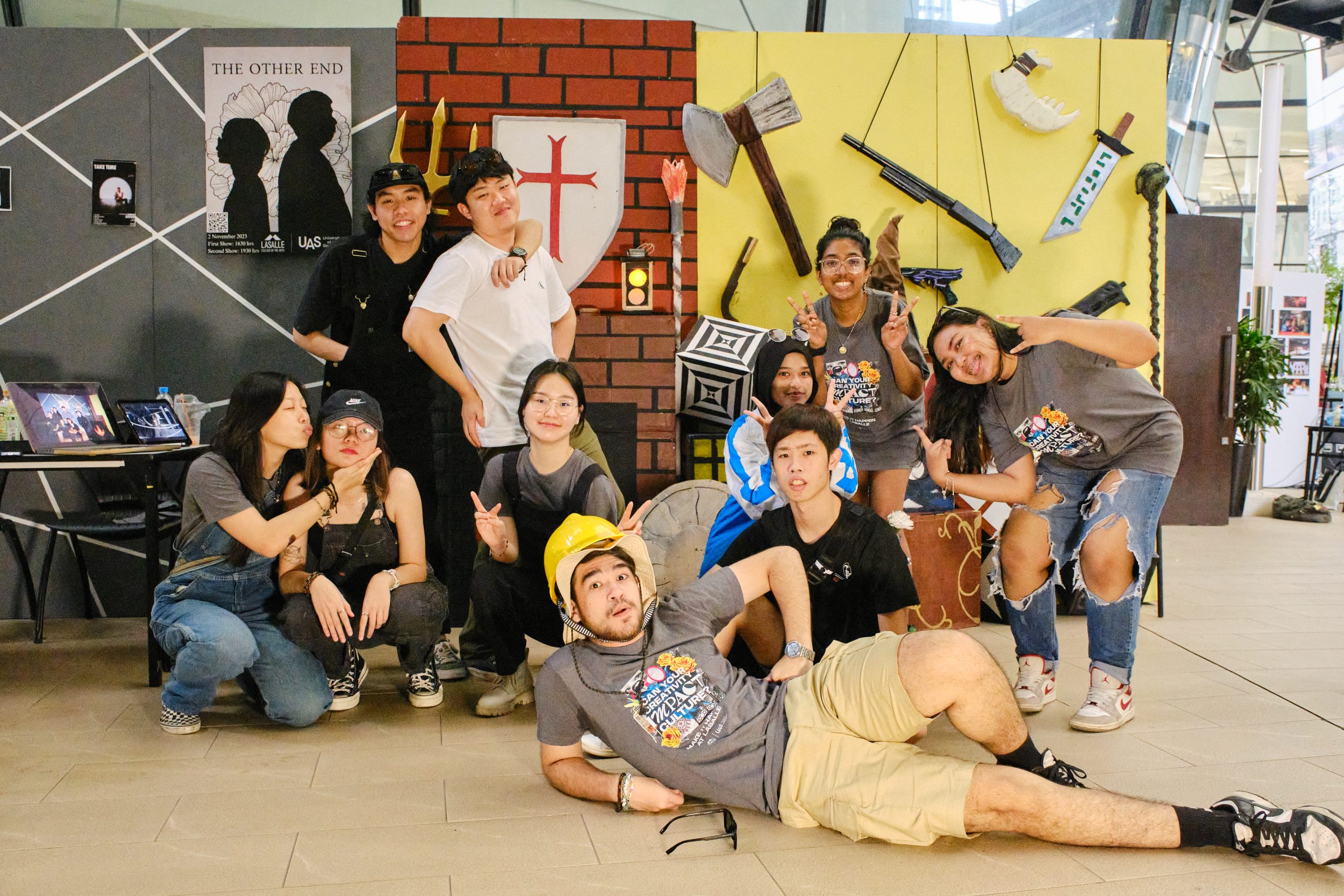 A group of students pose playfully in front of a wall decorated with medieval props and oversized weapons at an indoor campus event.