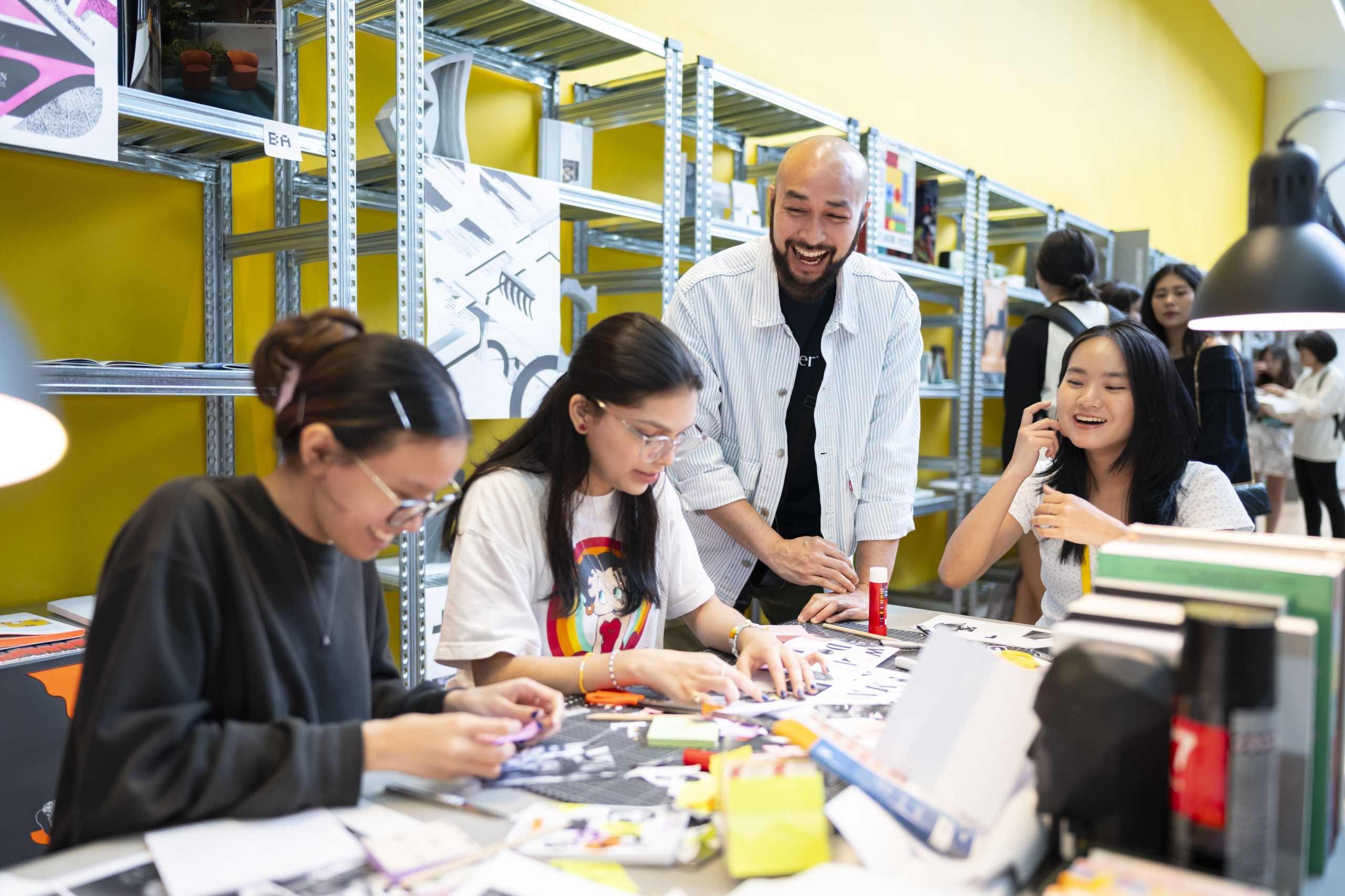 A LASALLE lecturer interacts with students working on design collages at a table, surrounded by bright artwork and studio shelves.