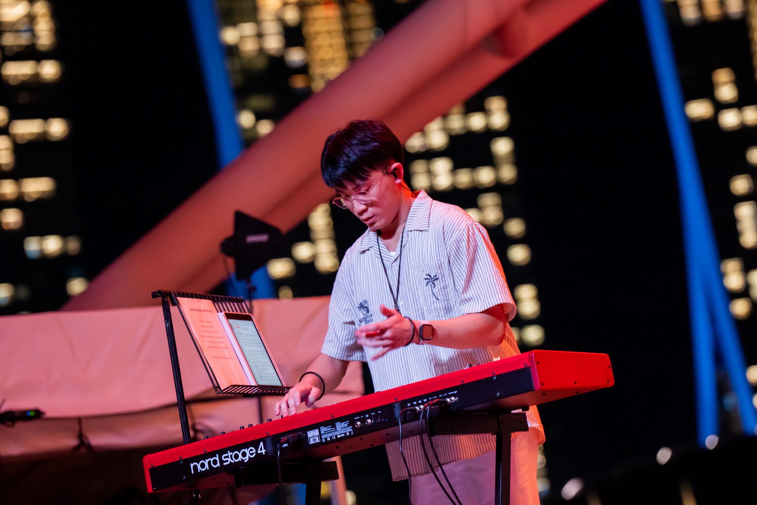 Performer playing a Nord Stage 4 keyboard on an outdoor stage at night, with music stand and city lights in the background.